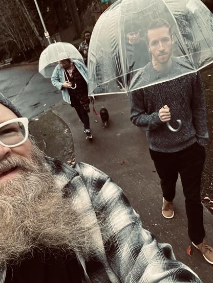 Selfie showing half of me with three people walking behind me. Two are taking refuge under umbrellas, one is walking a small black dog.