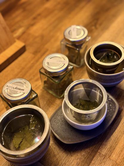 Preparing three small cups for a taste test - three small green tea cups have wire mesh baskets either leaves and hot water in them on a kitchen bench. The square glass jars with dried leaves inside and labels stuck to their silver lids sit behind - this was not a blind test.