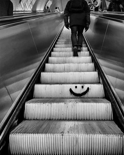 A person ascends an escalator, with a playful smiley face drawn on the steps. The scene is depicted in black and white.