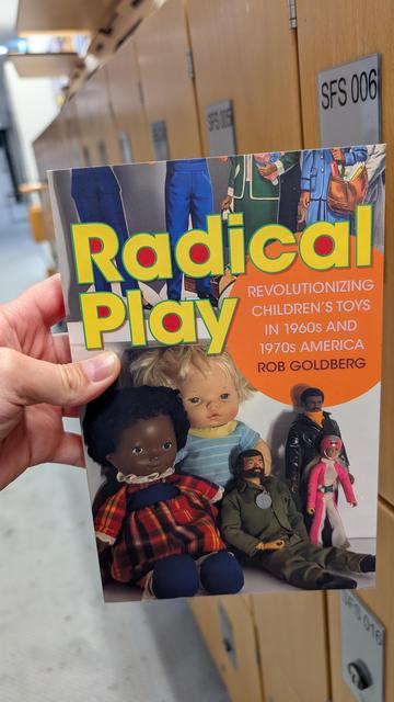 Holding the book "Radical Play" in front of a row of wooden lockers