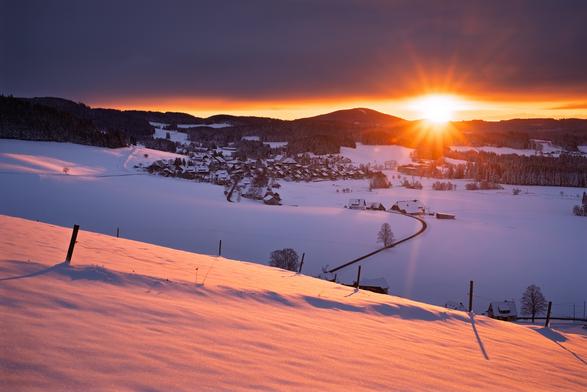 Sonnenaufgang über einer winterlichen Landschaft im Schwarzwald. Im Vordergrund eine tief verschneite Wiesenfläche mit einigen Zaunpfählen, die im Licht der aufgehenden Sonne rot leuchtet. Dahinter, etwas tiefer gelegen, eine offene Landschaft, die noch im Schatten liegt, mit dem Dorf Breitnau. Darüber eine bewaldete Hügelkette. Der Himmel ist bedeckt, abgesehen von einem schmalen Streifen direkt über dem Horizont, wo in der rechten Bildhälfte gerade die Sonne aufgeht.