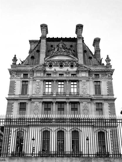 Photographie en noir et blanc de la pointe d'une aile du Louvre. Le ciel gris mais clair qui entoure le bâtiment réhausse la palette monochrome, comme le soleil qui se reflète dans les fenêtres centrales du second étage.