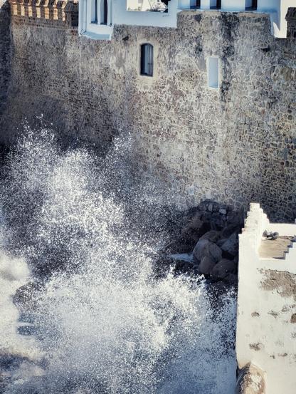 A vertical shot capturing a massive burst of white ocean spray crashing violently against the high, textured stone wall of an ancient coastal fortress. The wall features rough masonry, a dark recessed window, and white-washed sections near the top. To the right, on a lower white ledge, two pigeons perch calmly, seemingly undisturbed by the chaotic water spraying nearby