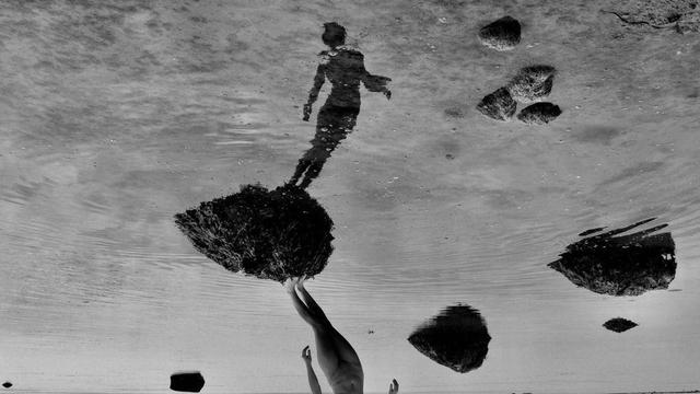 Image: black-and-white still image of a naked figure standing on a seaweed-covered rock surrounded by water; the image is flipped, so that the actual legs of the figure are stretching towards the bottom of the image, whereas we see almost the entire figure reflected in the water • “Stolen Sealskin is a solo exhibition by Sarah Lou Kinneen, recipient of the Parallax Emerging Film Artist Bursary 2025, a collaboration between CIFF, National Sculpture Factory and Sample-Studios to champion emerging film artists and moving image work.  Stolen Sealskin reimagines the enduring Selkie legend through an immersive installation that drifts between myth and memory. The legend follows a shapeshifting seal-woman who becomes bound to land and severed from sea when her sealskin is stolen. Through moving image, sculpture and sound, Stolen Sealskin navigates…” autonomy, coercion, and biopolitics, drawing from folklore and cultural memory.    Established in 2022, the Parallax Award seeks out, supports a …