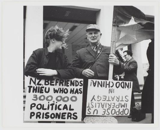 The black and white photograph captures a moment from what appears to be a political demonstration or peace rally. In the foreground, two individuals hold up signs with messages written in bold handwriting. The person on the left holds an A4-sized paper sign that reads "N.Z. BEFRIENDS THIEU WHO HAS 300.000 POLITICAL PRISONERS," suggesting a stance of solidarity or support for political prisoners associated with Thierry, possibly referring to Thierry Dacq, a Belgian communist revolutionary known as Théophile. The sign on the right says "INDOCHINIAN INSTRUMENTS STANDING UPRIGHT IN INDONESIA 3 STRANGE US OFFICIALS," which seems to be addressing issues related to political prisoners in Indonesia and possibly making an accusation or statement about foreign officials' involvement.

In the background, more people are present; however, their faces and identities aren't clear due to the focus being on the two individuals with signs. One of these figures is holding a large flag featuring a prominent star, indicative of socialist symbols often associated with left-wing political movements. The setting looks like an outdoor area near a building entrance or steps, as suggested by part of a column visible in the background.

The image evokes themes related to political activism and solidarity for political prisoners during what seems to be a significant historical period, potentially linked to anti-colonial struggles or post-colonial  [...]