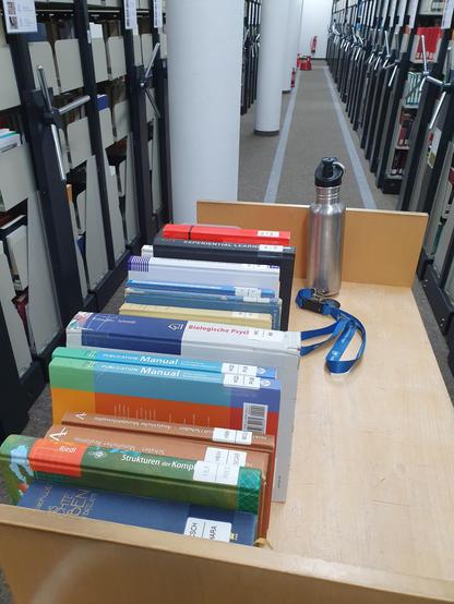 Bücherwagen im PSY-Bereich der UB. Darauf eine Reihe Bücher, eine Trinkflasche und ein Schlüssel.