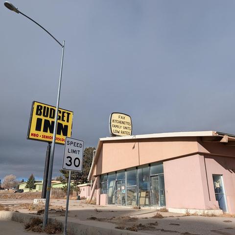 Photo of the Budget Inn, in Rawlins, WY. The peach building has unusual angles, accentuated by the perspective from the street, with grey clouds and blue sky peeking through. The large glass windows show the motel office front is being used for storage. A silver light pole stands just in front of a square, yellow sign for "Budget Inn", with "HBO" and "Senior Discount" in smaller text.  Above the roof of the building, an aging sign reads:
Fax 
Kitchenette
Family Units
Low Rates
