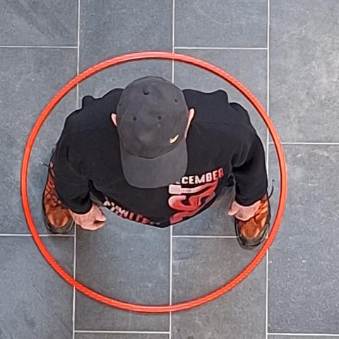 Blick von oben auf einen Mann mit schwarzer Kappe, schwarzem Sweater und rot-schwarzen Schuhen. Er steht in der Mitte eines roten Hula-Hoop-Reifens. Der Boden besteht aus großen, graumelierten Kacheln und schmalen weißen Fugen.