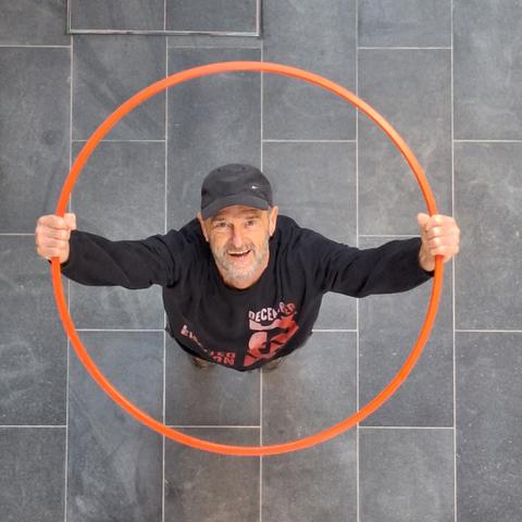 Wieder der Blick von oben. Der alte Mann mit grauem Bart hält den Hula-Hoop-Reifen über seinen Kopf und blickt dabei nach oben.