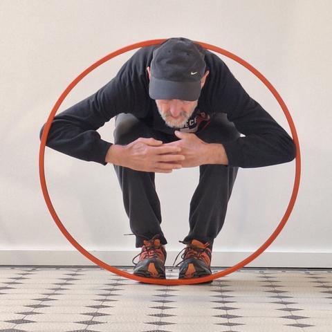 Diesmal kommt der Blick frontal von vorne. Innerhalb des aufrecht stehenden Hula-Hoop-Reifens steht der alte Mann nach vorne gebeugt. Die Arme sind angewinkelt, die Hände beieinander. Der gebeugte Körper befindet sich komplett innerhalb des Reifens. Im Hintergrund eine weiße Wand mit weißen Fußleisten. Der Boden besteht aus weißen Fliesen mit kleinen schwarzen Rauten.