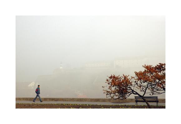 A horizontal photo taken on a foggy day showing a person walking along a paved path with a low stone wall on the right side. The person is walking right and is wearing a blue jacket with a red stripe across and jeans. To the right of the path is a bench next to a small tree with reddish-brown autumn leaves. The background is obscured by dense fog, but a fortress with a white clocktower is faintly visible on the distant hill.