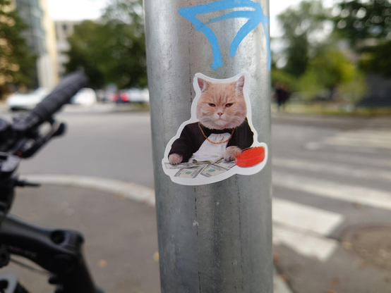 A sticker on a metal pole shows a fluffy cat in a black suit and gold chain sitting behind a pile of dollar bills and a small bowl of caviar.