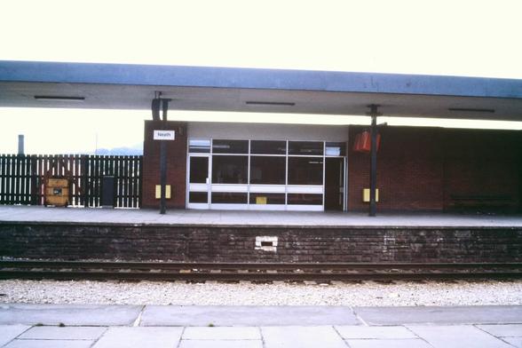 Foto im Querformat. Vom gegenüberliegenden Bahnsteig aus blickt die Kamera auf ein einfaches, einstöckiges Bahnhofsgebäude aus rötlich-braunen Backsteinen, das eine mehrteilige Fensterfront in voller Höhe zur Wartehalle hat. Über dem Bahnsteig ist ein flaches Dach, das von Metallträgern getragen wird. An einem dieser Träger, halb links, hängt das Bahnhofsschild mit der Aufschrift „Neath“. Links neben dem Gebäude ist ein einfacher Holzzaun.