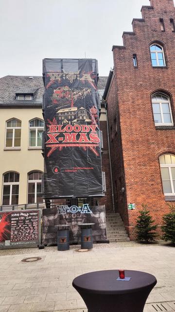 Auf dem Campus Mitte der Charité ist eine Feuerleiter in ein Banner gehüllt, welches das Logo vom W:O:A (Wacken) und dem Bloody X-Mas Market hüllt. Darunter steht das Logo des Zentrums für Transfusionsmedizin und Zelltherapie Berlin (ZTB). Darunter prangt ein WOA Logo aus LEDs und zwei Stehtische. Im Vordergrund befindet sich ein Stehtisch mit schwarzer Husse. Im Hintergrund neben der Feuerleiter ist ein rotes Backsteingebäude mit ein paar kleinen Tannen davor.