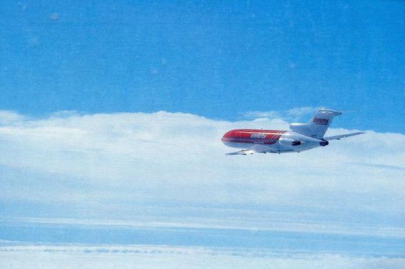 A bright red-and-white Emery Worldwide Boeing 727 soars high above a blanket of soft, brilliant white clouds. The tri-engine jet is captured in a graceful right-bank turn, its distinctive T-tail and three rear-mounted engines clearly visible. The aircraft’s fuselage carries bold EMERY titles in large white letters over a vivid red forward section that fades into clean white toward the tail. The sky above is a deep, uninterrupted blue, creating a striking contrast with the aircraft’s colors and the cloud layer below. The scene conveys both motion and altitude, giving a sense of high-level cargo operations during the classic era of 727 freighters.
