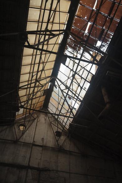 Looking up at a long row of stained, weathered skylight windows at the centre of a pitched roof of a disused industrial building - a dark metal framework silhouetted against the light filtering through the dirty glass. Away from and below the skylights, a grimy brown darkness.