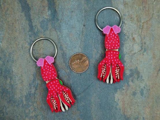 Two 3d printed Strawberry Squid designs. The Strawberry Squid has a red body covered with pinkish specs and pinkish fins on the mantle. The eye on the left is small and dark blue, the eye on the right is large and greenish-yellow. The tentacles and suckers on the arms are tan. Each squid has a 1-inch (2.5cm) spit ring though the top of the mantle. Between them is a US penny for scale.