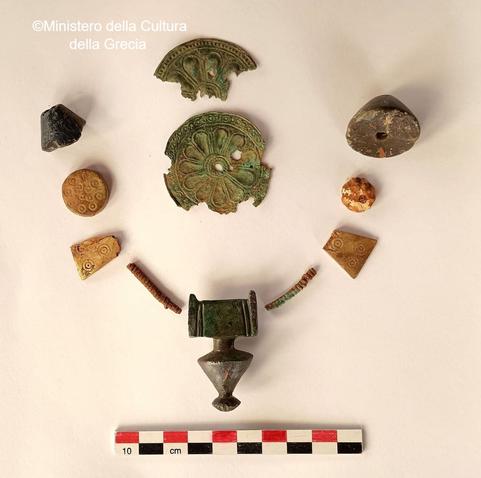 Reperti archeologici in metallo e ceramica, tra cui un anfora, frammenti decorativi e altri oggetti, disposti su un fondo bianco con un righello per scala.