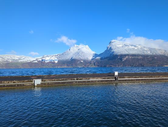 Bild von einem blauen stillen See. Der Himmel ist Strahlend Blau. Im Vordergrund ist eine Mohle. Auf der anderen seite des Sees ist eine Bergsichel zu sehen. Die Oberen zwei drittel der Berge sind schneebedekt. Auf einer Klaren kante wechselt es zu grünem Wald. Dies ist die Schneefallgrenze letzter Nacht