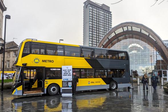 A modern, black and yellow double-decker bus, branded 'METRO', is parked outside the distinctive curved, glass-roofed entrance of the Sheffield Interchange, with a tall city building in the background. A man in a uniform stands next to a promotional banner advertising a survey about buses. The ground is wet and reflects the bus. Photo by Evie Carroll, South Yorkshire Mayoral Combined Authority.