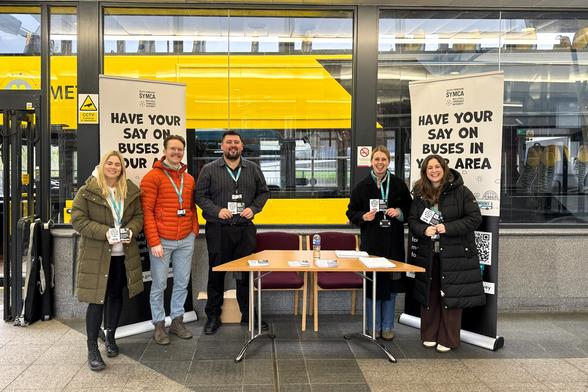 Five people, three women and two men, stand behind a small folding table indoors at a public transport hub. Two large black and white banners on either side read, 'SYMCA HAVE YOUR SAY ON BUSES IN YOUR AREA'. They are holding information leaflets, and a yellow double-decker bus is visible through the glass windows behind them. Photo by Evie Carroll, South Yorkshire Mayoral Combined Authority.