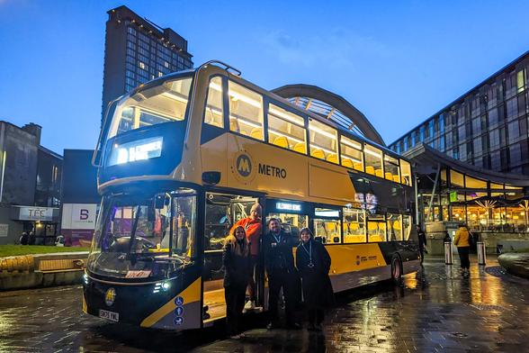 A modern, yellow and black 'METRO' branded double-decker bus is parked outside a city building at dusk. The bus interior lights are on, illuminating the upper deck seats. Four people are standing on the steps by the open front door of the bus. The foreground is wet, reflecting the light. Photo by Evie Carroll, South Yorkshire Mayoral Combined Authority.
