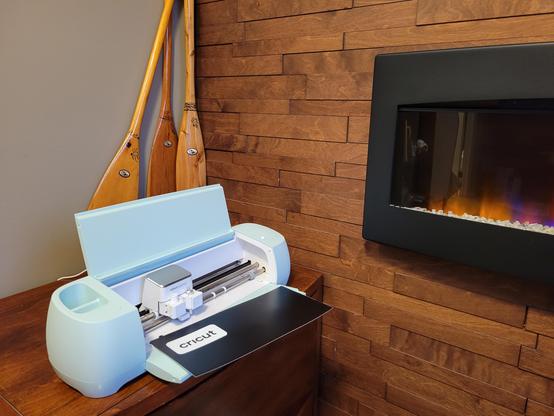 A mint-coloured Cricut vinyl cutter sits on a cabinet next to an electric fireplace, with three child-sized paddles behind it.