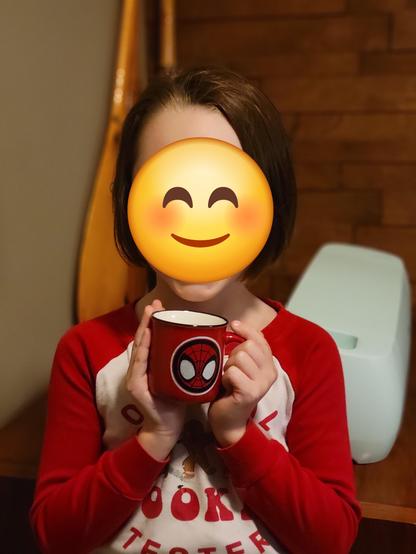 A kindergartener in pyjamas holds a tiny mug with a stylized Spidey and Friends graphic applied to it. Her face is hidden by a smiley emoji.