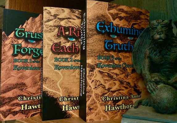 The three books in the Kovenlore series, out of seven, sit on a bookshelf, a gargoyle bookstand on the right.

Each book’s cover is the negative image of a golden, hand drawn map showing mountains, plains, and rivers, but sometimes a black void. The titles are always displayed across two lines up high, the color varying. Between the two lines of the title is always a set of turquoise eyes.

A subtitle, always in turquoise, is below that and shares which number book it is in The Kovenlore Chronicles. At the bottom is the author’s name, Christina Anne Hawthorne, and always in a dark yellow. All text is outlined in black, the titles possessing a shadow.

Book 1, Trust in the Forgotten, is on the left, it’s title in turquoise. Book 2, A River in Each Hand, is at center, its title in red. Book 3, Exhuming Truths, is on the right, its title in dark blue.