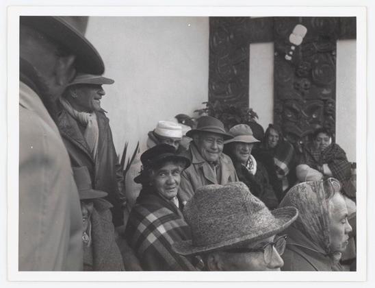 The photo captures a group of people gathered closely together, possibly at an outdoor event or gathering. The setting seems informal and social with individuals wearing various types of hats indicative of cold weather attire, such as fedoras and wide-brimmed hats for sun protection. Some wear overcoats, scarves, and shawls suggesting the photograph was taken in a temperate climate during cooler seasons like fall or early spring.

The group is diverse; men and women are present with both younger-looking individuals alongside those who appear older. The expressions on their faces range from smiles to neutral looks, indicating varying levels of engagement with what's happening around them. One individual stands out as they wear glasses and a patterned scarf wrapped snugly against the chill.

The setting includes architectural elements like carved wooden panels in the background which might suggest cultural significance or simply be part of local aesthetics. The absence of modern technology hints at an earlier time period, possibly mid-20th century given the style of clothing and overall ambiance of the photograph.

It is important to note that this description relies on visual analysis without additional context provided by external sources like Wanganui - Marae links which are not clickable in my current function. The image could be from various regions where such gatherings occur, but a specific location or event isn't c [...]