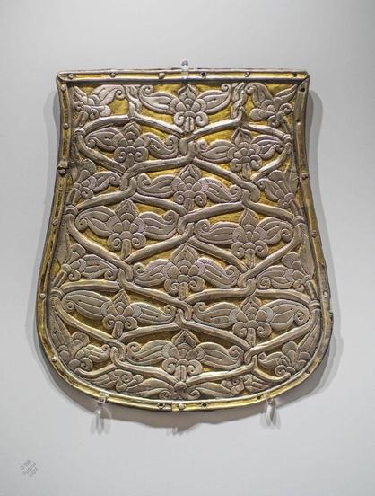 A photograph of the object on display in museum mounts and lighting. It's a vaguely tongue-shaped piece, silver with selective gilding (or maybe just wear). The decoration is an islamic-looking allover repeat pattern of twisted cord net with a leaf and flower motif, possible derived from a textile pattern. It's dated to CE 900-930, a time before the Hungarian state existed in a recognisable form, when the Carpathian basin was being occupied by a coalition of tribes that claimed direct descent from Atilla the Hun, defeating various early European / late Roman armies claiming it. It's not known where the object originated but stylistically it's consistent with Samanid/Sogdian design.