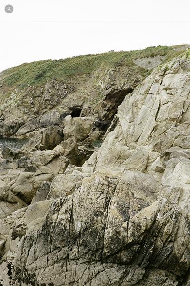 Klippenküste mit großen, hellen Felsen im Vordergrund; dahinter grasbewachsene Hänge und kleine Felsnischen unter bewölktem Himmel.