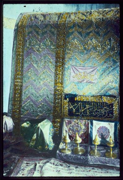 The image depicts a richly decorated religious shrine, featuring intricate patterns and ornate calligraphy. The dominant colors are golds, blues, greens, and purples with embossed textures that suggest the use of metal leaf or similar materials for embellishment.

At the top center hangs a rectangular banner in white fabric adorned with Arabic script, possibly indicating its sacred significance. Below this, on what appears to be an altar-like platform, there are two small golden-colored bowls supported by slender stands, alongside another item reflecting light which could suggest it is made of metal or glass. 

The background showcases the shrine's intricate designs: green and purple chevron patterns that frame a central field with Arabic calligraphy in gold hues. The script likely contains verses from the Quran or other religious texts, serving as blessings or prayers for those who visit.

To the left side on the ground lies an object partially obscured by another layer of fabric; its details are not clear due to this partial obstruction and angle at which it's presented.

Below all these items is a patterned rug with floral motifs in muted colors like pinkish-purple, beige, green. The lower portion also reveals more draped fabrics or garments contributing to the overall sacred ambiance of the scene.

This photograph was likely taken during an exploration related to biblical sites, as suggested by references such as 'Ro [...]