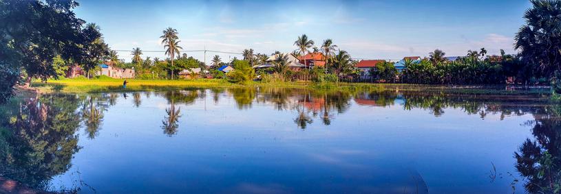Photo shows a deep blue oval lake framed by trees and with small houses of a local village in the background. A lone fisher is testing his fishing rod and fishing luck on the far side of the lake. It is early morning after sunrise, and the sun is shining from a partly clouded, blue sky. Cambodia is as safe, comfortable and kind as ever. Angkor and Siem Reap welcomes visitors.