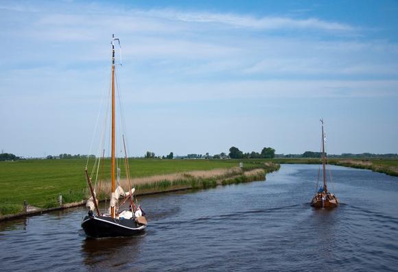 Twee traditionele zeilboten op een smal kanaal in een Nederlands landschap. De voorste boot vaart naar links, met een groot houten zeil en lichte tuigage. Op het dek liggen enkele voorwerpen en er zitten mensen op de boot. Aan de rechterkant, verderop op het water, is een tweede, kleinere zeilboot te zien met ook enkele mensen erop. Aan weerszijden van het kanaal liggen groene weilanden met lage begroeiing. De lucht is blauw met wat lichte wolken. Het geheel heeft een rustige, landelijke sfeer.