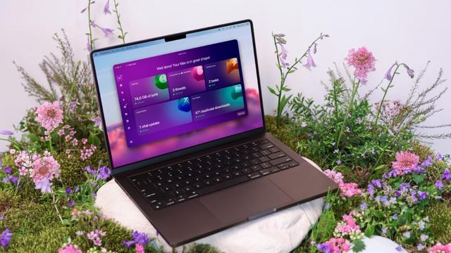 A MacBook Pro resting on a white stone surrounded by fresh grass and pink wildflowers, displaying the new CleanMyMac Smart Care dashboard on the screen with scan results showing "74.6 GB of junk" and "3 threats" found.