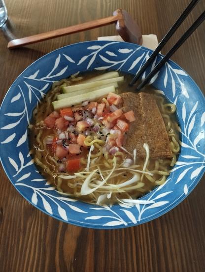 Un primer plano de un tazón de sopa de fideos (ramen) sobre una mesa de madera. El tazón es azul con un patrón de hojas blancas y contiene fideos amarillos en un caldo claro. Los toppings incluyen dados de tomate rojo, cebolla morada, y semillas de sésamo negro. También se ven tiras rectangulares de un ingrediente marrón (posiblemente tofu o proteína vegetal), rodajas finas de pepino verde pálido y tiras rizadas de cebolla blanca o cebolleta. Un par de palillos negros están apoyados en el borde del tazón. En el fondo, una cuchara de madera de mango largo descansa sobre la mesa de madera oscura.