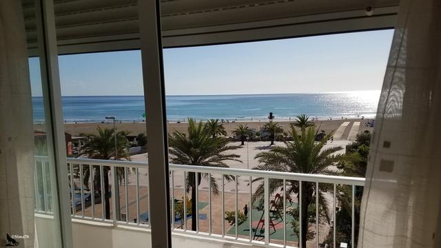 Vue sur mer depuis porte-fenêtre donnant sur une promenade avec palmiers et plage.
Sea view from a French window overlooking a promenade with palm trees and a beach.
Vista al mar desde una puerta-ventana que da a un paseo con palmeras y playa.
