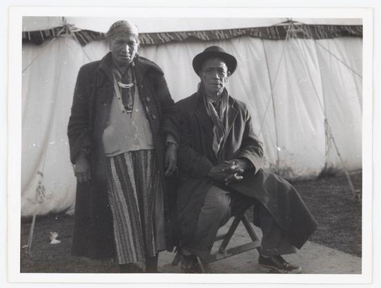 This black and white photograph features two individuals standing close together against a backdrop of what appears to be temporary, canvas tents. The person on the left is wearing traditional or ceremonial attire that includes a striped skirt-like garment with vertical patterns in dark colors, possibly indicative of Native American culture. They are adorned with several necklaces and other accessories which suggest cultural significance. Their expression is neutral but carries an air of dignity.

The individual on the right seems to be dressed more informally yet still holds themselves firmly within what appears to be a traditional context as well; they wear a hat, coat, scarf or shawl draped around their shoulders, and possibly gloves underneath the coat. The setting suggests this photograph was taken at an outdoor event that involves temporary structures such as tents often associated with gatherings like fairs, carnivals, or cultural festivals.

The background is out of focus but shows ropes supporting the canvas tent structure which gives context to the environment in which these individuals are situated. There's a sense of historical significance given by both their attire and the black-and-white medium used for this photo.