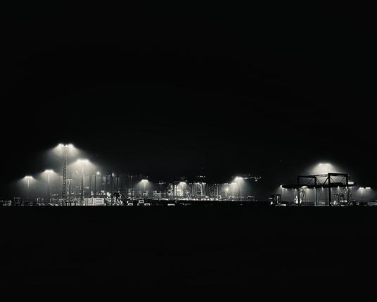 The image shows a night-time view of Container Terminal IV in Bremerhaven, captured from a distance so that the facility appears as a thin, glowing band in an otherwise pitch-black landscape. Powerful floodlights illuminate the cranes, container stacks and port structures, creating misty halos that cut through the darkness. The industrial scene feels both massive and strangely quiet, as if the entire terminal is suspended in a sea of night, defined only by scattered islands of artificial light.