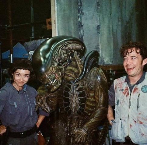 Photographie en plan taille de trois protagonistes de la série Alien Earth.
A gauche, l'héroïne principale, une jeune fille, cheveu noir, souriante se tenant les mains sur la taille et faisant la pose.
Au centre la créature alien monstrueuse avec son exosquelette noir et lustré à la manière d'une écrevisse.
A droite, le héros masculin qui joue le frère.
Son regard est tournée vers la jeune fille à sa droite et il semble effrayé par la présence de la créature tout en étant souriant devant le sourire de sa sœur.
Sentiments mêlés.