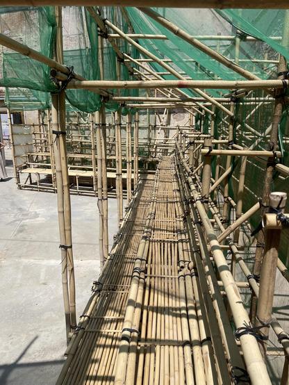A large matrix of bamboo sticks, formed into scaffolding, viewed from the inside. The structure is covered in green netting, situated in a Venetian courtyard.
