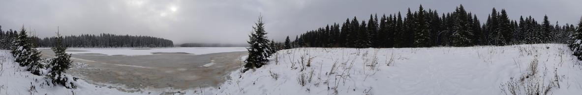 A panorama photo taken from the shore of a partly frozen lake. The shore os covered in snow, the forest is in the distance. The weather is gray. the sin barely makes it through the cloud cover.