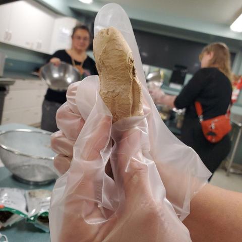 Close-up of a person's gloved hand holding a piece of dried plant root, possibly ginger, wrapped in a clear plastic bag, with two other people and kitchen equipment in the blurry background.