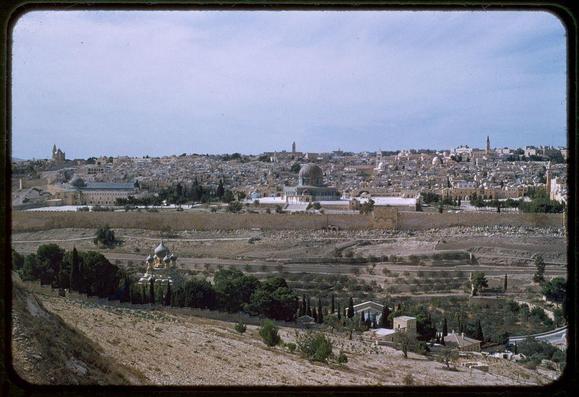 The image depicts a wide, panoramic view of an ancient city with dense clusters of buildings spread across the landscape. The foreground is dominated by barren fields and patches of vegetation on undulating terrain that appears arid or semi-arid.

In the mid-ground, various religious structures are visible: prominent among them is a large white-domed building which stands out due to its size compared to other surrounding architecture. Its structure suggests it could be an important cultural or historical site, possibly related to major religions given such domes' common symbolic association with sacred spaces in Islamic and Christian faiths.

Additionally, there are smaller religious structures, including one that resembles a traditional Eastern Orthodox church with distinctive onion-shaped cupolas, indicating the presence of diverse religious communities. 

The background reveals more densely packed buildings, hinting at an urban sprawl extending towards the horizon under a clear sky. The overall impression is that of a historically rich and culturally complex cityscape, likely Jerusalem given the historical context provided by "Jerusalem from Bethany Road between 1948 and 1958."