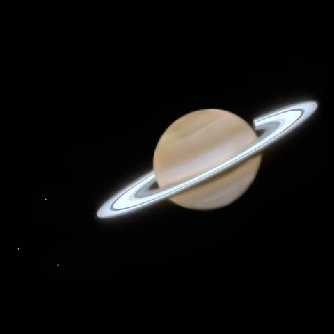 In this JWST image, Saturn appears as a pale-gold sphere with faint horizontal bands, encircled by a bright, thin oval ring. Three tiny white points to the lower left represent the satellites Dione, Enceladus and Tethys against the black background.