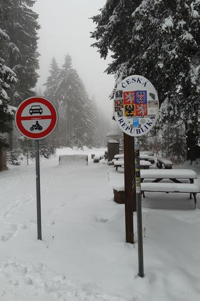 Photo of the border crossing from Germany into the Czech Republic. The photo shows a snowy forest track with several foot tracks. There is a sign "Česká republika", covers with some german stickers. Next to it is a czech traffic sign meaning cars and motorcycles are prohibited. There are a few snow-covered benches on the czech side.