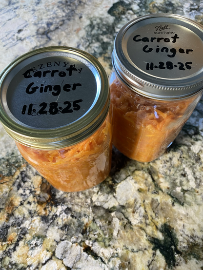 We see two clear jars from 3/4 above. The silver lids say “Carrot Ginger 11-28-25.” We see shredded carrots in vibrant orange.