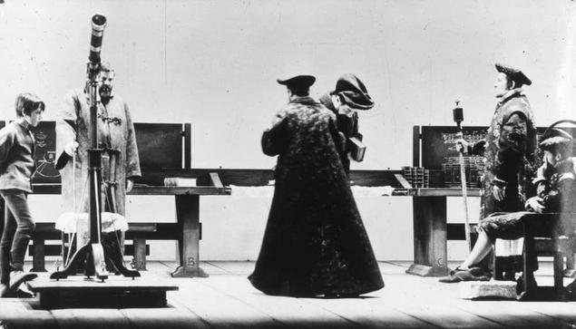 The image is a black and white photograph depicting four individuals engaged in what appears to be an astronomical or scientific observation. The setting seems historical, as suggested by the attire of the figures which includes hats, coats, and gloves typical of past eras.

On the left side stands a young boy holding a large telescope mounted on a tripod stand, suggesting he may have been observing celestial objects at that time. Opposite him, two men are seated behind tables with various scientific instruments including what looks like an armillary sphere and other measuring devices which could be used for navigation or astronomy.

In the center of the image stands another man in dark attire engaged deeply as if inspecting something on a table covered by a black cloth. His posture indicates focus and importance, hinting he might be analyzing data or findings from their observations.

To his right is an elderly figure with a long beard wearing spectacles and seated at what seems to be a writing desk. This person appears contemplative, possibly reflecting upon the results of their work.

The background features several wooden benches and tables arranged in rows which may indicate that this gathering was part of some scientific demonstration or public lecture related to astronomy or navigational sciences. The overall atmosphere is one of seriousness and intellectual pursuit.
