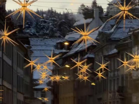 Eine malerische Straße, die mit leuchtenden sternförmigen Lichtern geschmückt ist und eine festliche Atmosphäre schafft. Schneebedeckte Dächer und eine bergige Kulisse werten die winterliche Atmosphäre auf.